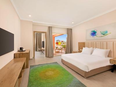 Modernes Hotelzimmer mit großem Bett, grünem Teppich, Fernseher und Balkon mit Meerblick.