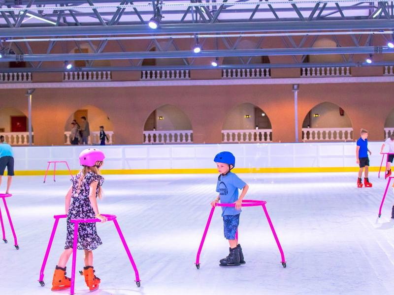 Kinder beim Eislaufen in einer Halle mit pinkfarbenen Gehhilfen auf dem Eis.