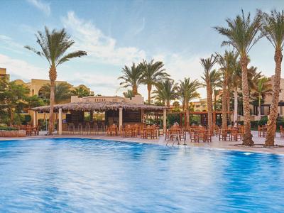 Poolbereich eines Resorts mit Palmen, Liegestühlen und Sonnenschirmen unter blauem Himmel.