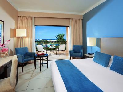 Bright hotel room with blue accents, double bed, and balcony overlooking the sea.