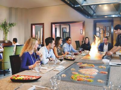 Guests sit at a large table in a restaurant as a chef prepares dishes.