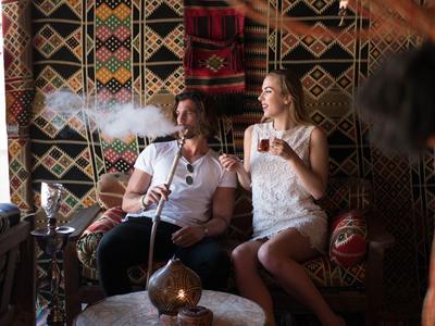 Dos personas descansan en una habitación tradicional con alfombras estampadas y pipa de agua.