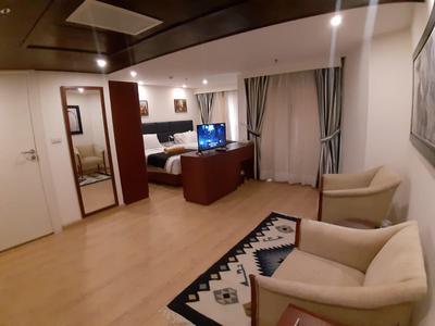 Ruime hotelkamer met bed, tv, fauteuils en een patroon tapijt op houten vloer.