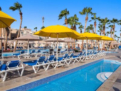 Pool area with blue loungers and yellow umbrellas near palm trees and buildings under clear sky.