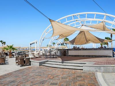 Sunlit outdoor seating area with chairs and tables under a shaded structure on a clear day.