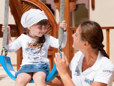 Mädchen auf einer Schaukel, betreut von einer Frau in einem Hotelumfeld.