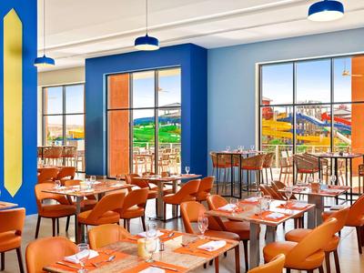 Helles Restaurant mit blauen Wänden, großen Fenstern und orangen Stühlen an Holztischen.