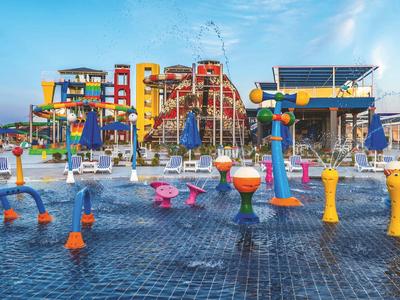 Kleurrijk waterspeelgebied met sproeiers voor een hotelcomplex bij een heldere lucht.