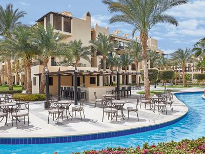 Außenbereich eines Hotels mit Pool, Tischgarnituren und Palmen unter blauem Himmel.