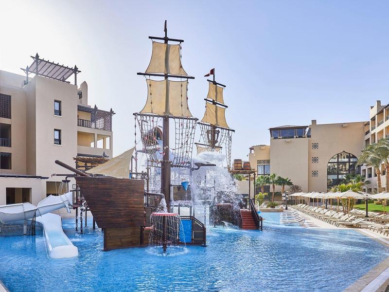 Parque acuático con barco pirata, toboganes y piscina en un resort hotelero bajo cielo despejado.