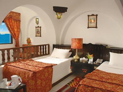 Habitación de hotel acogedora con dos camas individuales, cobijas rojo-doradas y vista al mar.