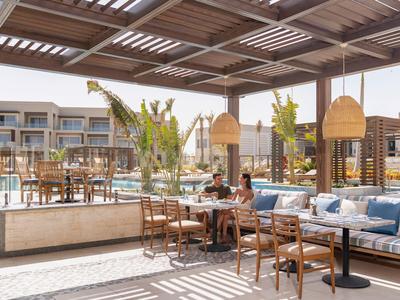 Überdachte Terrasse mit Sitzbereich und Blick auf modernes Hotelgebäude und Pool.