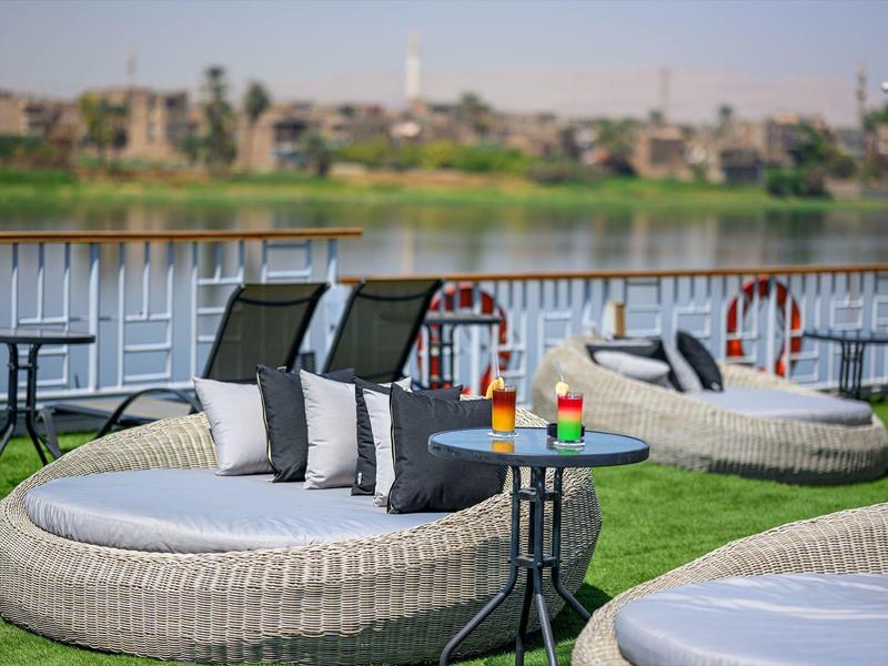 Área lounge con sillones de ratán y cócteles coloridos junto a un río con vista a la ciudad al fondo.