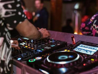 DJ bedient bunte Mischpult- und Plattenspieler in dunkler Club-Atmosphäre mit unscharfem Publikum.