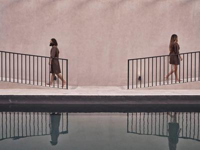 Two women walk along a walkway by a pink wall with a pool in the foreground.