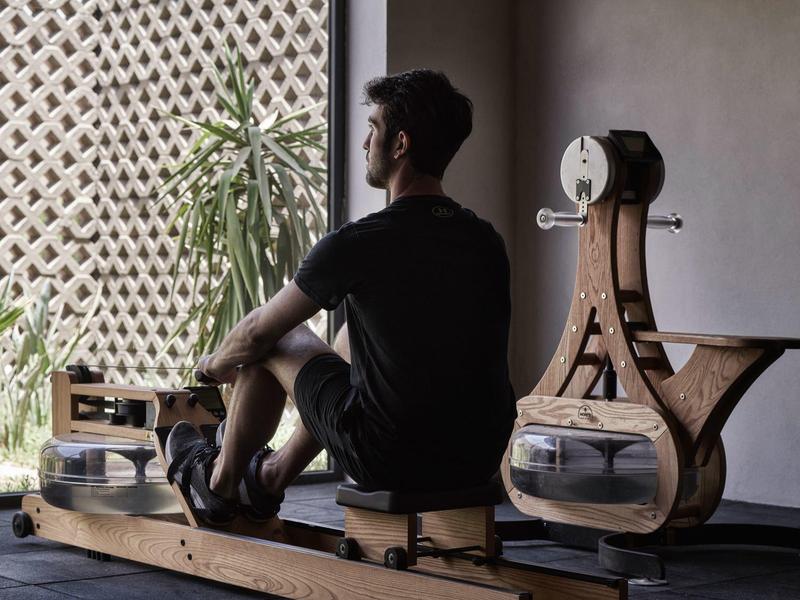 Man sits on rowing machine in modern, minimalist interior with plants and natural light.