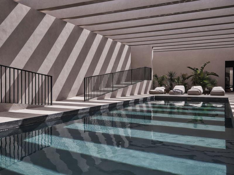 Modern indoor pool with sun loungers and striped shadows cast by beams on the wall.
