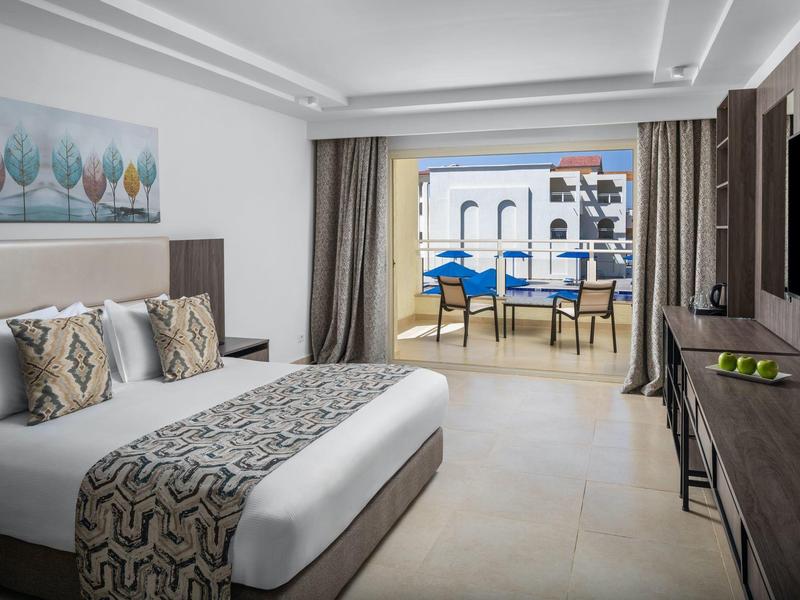 Helles Hotelzimmer mit großem Bett, Schreibtisch und Blick auf Balkon mit Tischen und Stühlen.