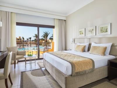 Chambre d'hôtel lumineuse avec lit double et vue sur piscine et palmiers.