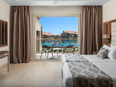 Chambre d'hôtel moderne avec balcon donnant sur une piscine et des bâtiments sous un ciel bleu.