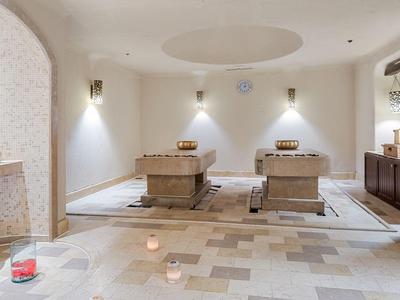 A serene spa room with two massage tables, soft lighting, candles, and beige tiled flooring.