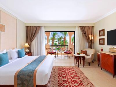 Bright hotel room with large bed, seating area, and balcony overlooking tropical plants.