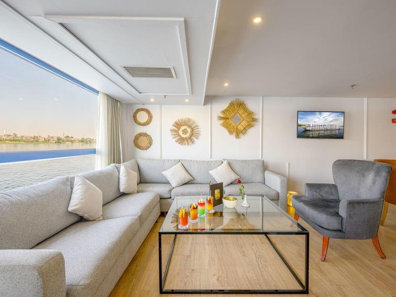 Bright hotel lounge with beige sofas, armchair, glass coffee table, and large window with water view.