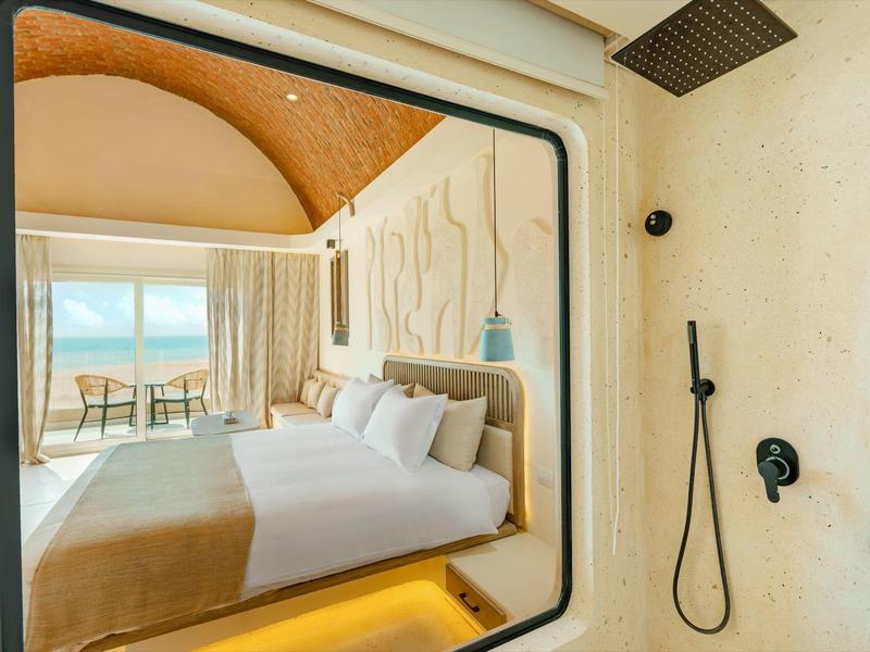 Moderne hotelkamer met groot bed, balkon met zeezicht en open douchegedeelte.