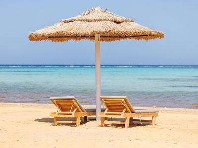 Two wooden lounge chairs under a straw beach umbrella on a sandy shore by clear blue sea.