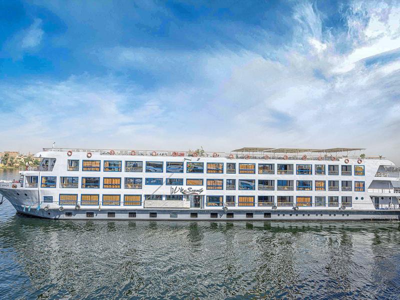 Large white river cruise ship with multiple decks floating on calm water under a blue sky.