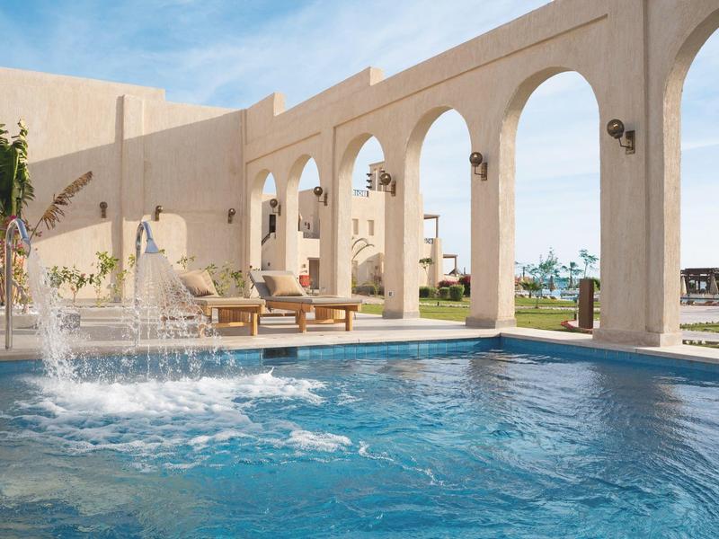 Outdoor pool with water fountain and arches in a sunny hotel courtyard.