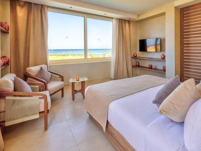 Chambre d'hôtel lumineuse avec grande fenêtre donnant sur l'océan, lit double, fauteuils et TV.