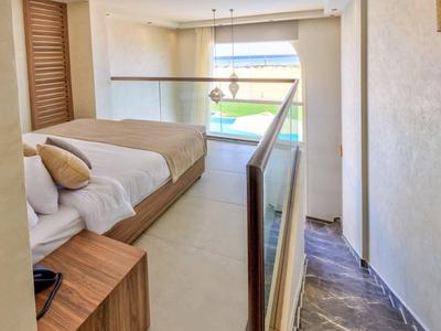 Chambre lumineuse avec un grand lit, mobilier en bois, rampe en verre, et vue sur une piscine extérieure.