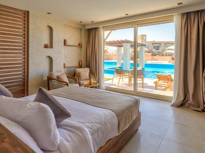Chambre d'hôtel avec grand lit, coin salon et portes coulissantes donnant sur une piscine et des transats.