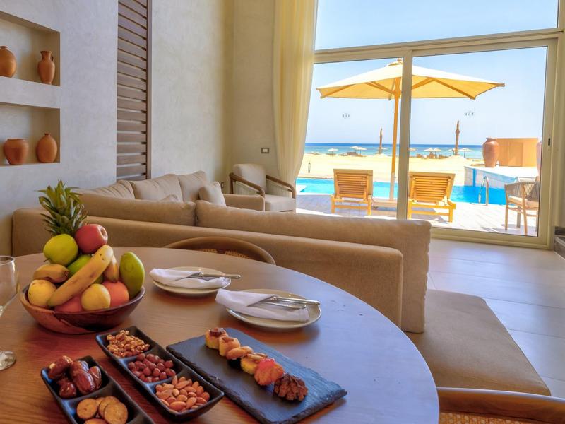 Coin salon confortable avec fruits frais et snacks, vue sur piscine ensoleillée avec parasols et chaises longues.