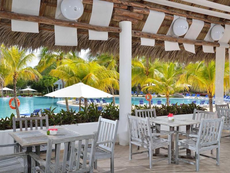 Zastřešená terasa s bílými stoly, výhled na bazén obklopený palmami.