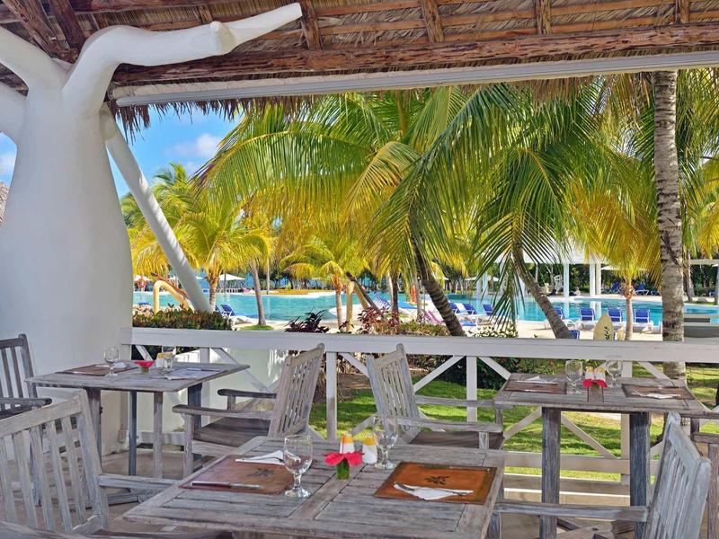 Zastíněná terasa s dřevěnými stoly a židlemi pod palmami u pláže.