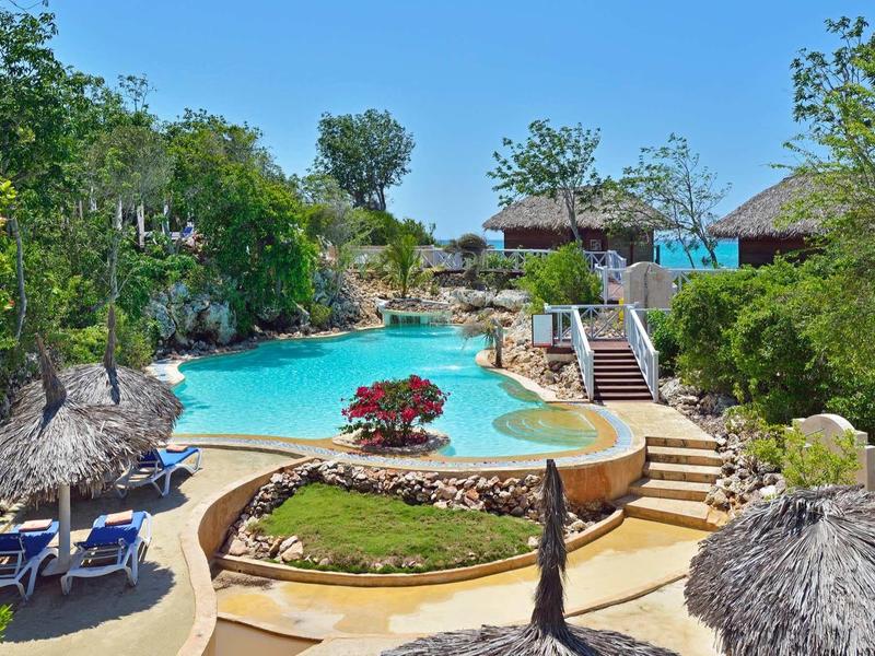 Venkovní bazén obklopený bujnou zelení a slaměnými slunečníky.