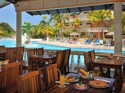 Ein schattiger Essbereich mit Holztischen und Blick auf einen Pool und Palmen.