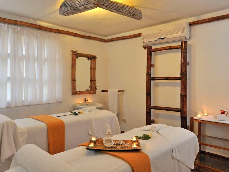 Gemütlicher Massageraum mit zwei Liegen, Holzmöbeln, Kerzenlicht und weißen Vorhängen.