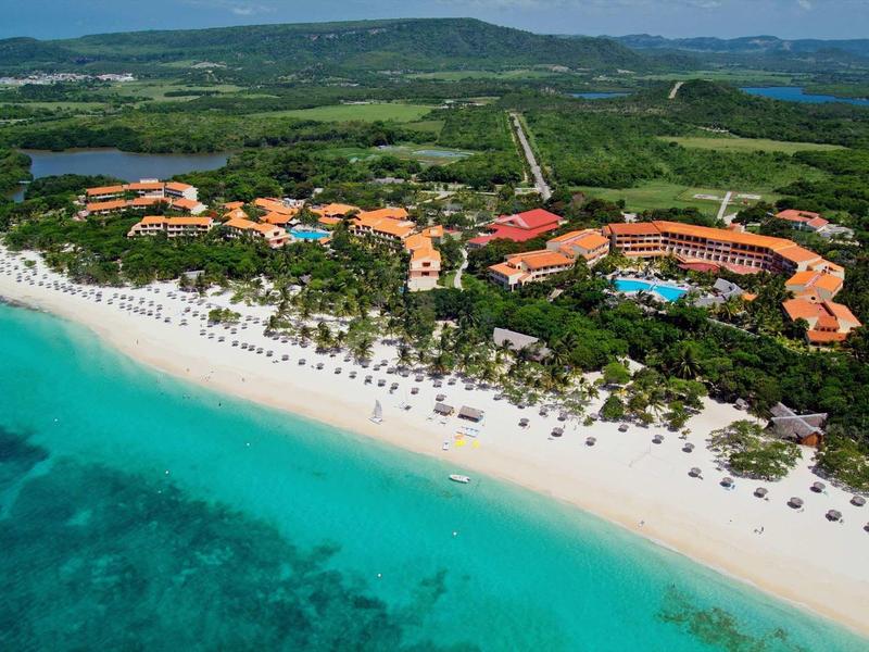 Langer weißer Sandstrand mit türkisblauem Meer, dahinter grüne Vegetation und große Ferienanlage.