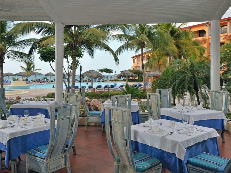 Offenes Restaurant mit blauen Tischdecken, Stühlen und Blick auf Strand mit Palmen und Sonnenschirmen.