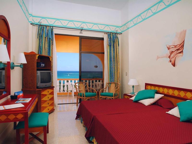 Hotelzimmer mit zwei roten Betten, grünem Stuhl, Holzmöbeln und Meerblick durch Fenster mit Vorhängen.