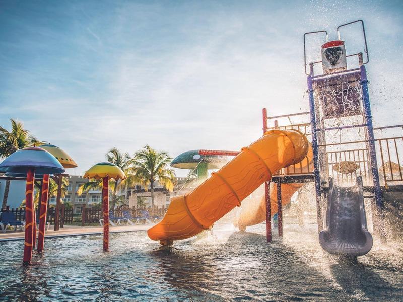 Dětské vodní hřiště s skluzavkami a barevnými slunečníky v hotelovém bazénu za slunečného dne.