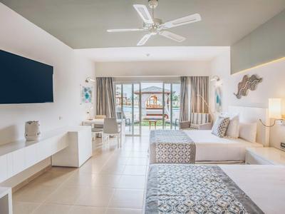 Helles Hotelzimmer mit zwei Betten, Flachbildschirm, Ventilator und Blick auf Terrasse mit Meer.