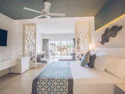 Modernes Hotelzimmer mit großem Bett, hellen Wänden, Deckenventilator und Balkon mit Meerblick.