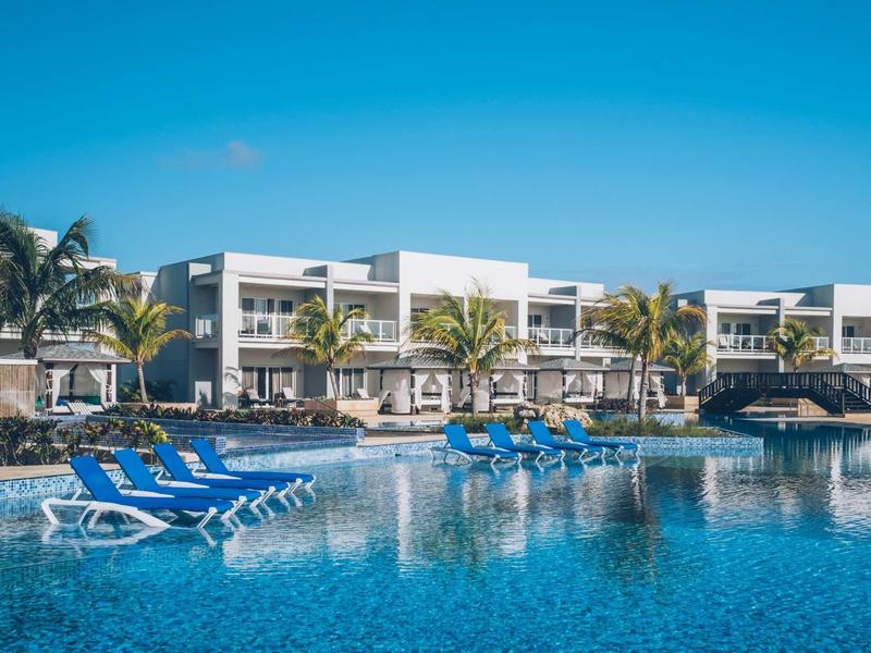 Moderner Hotelkomplex mit Pool, Sonnenliegen im Wasser und Palmen unter blauem Himmel.