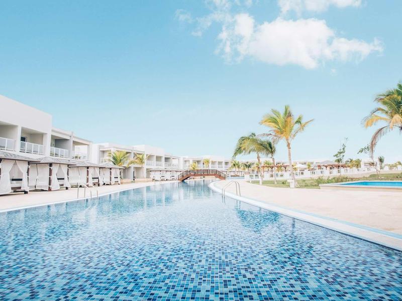 Großer blauer Pool mit Sonnenschirmen, Liegen, Palmen und weißem Gebäude im Hintergrund.