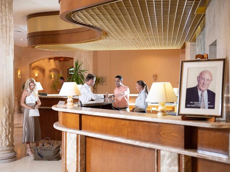 Recepción de hotel con tres huéspedes y un empleado, lámparas elegantes y una gran columna al fondo.