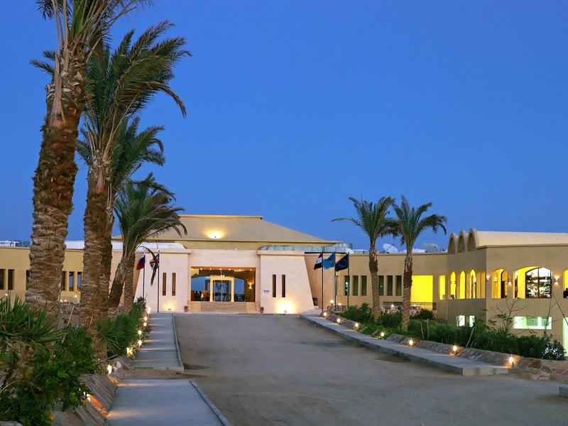 Edificio de hotel iluminado al anochecer con palmeras y camino de entrada amplio.